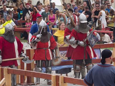 Cavalieri in armatura si sfidano in un evento medievale, con spettatori in abiti storici tutt’intorno.